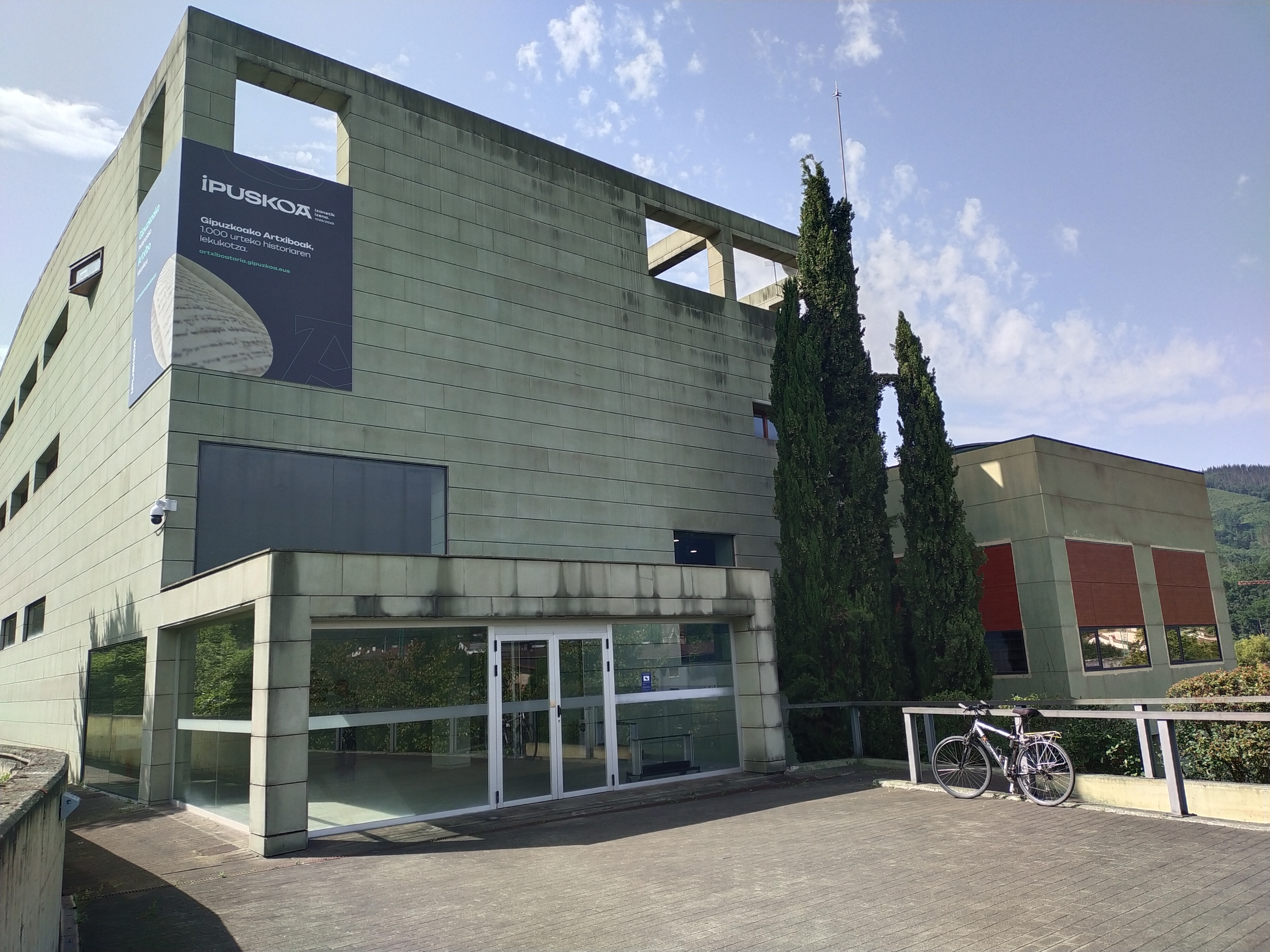 Entrada del Archivo Histórico Provincial de Guipúzcoa en Oñati: volumen de fachada verde-gris con aperturas geométricas y acceso acristalado bajo un pórtico voladizo.